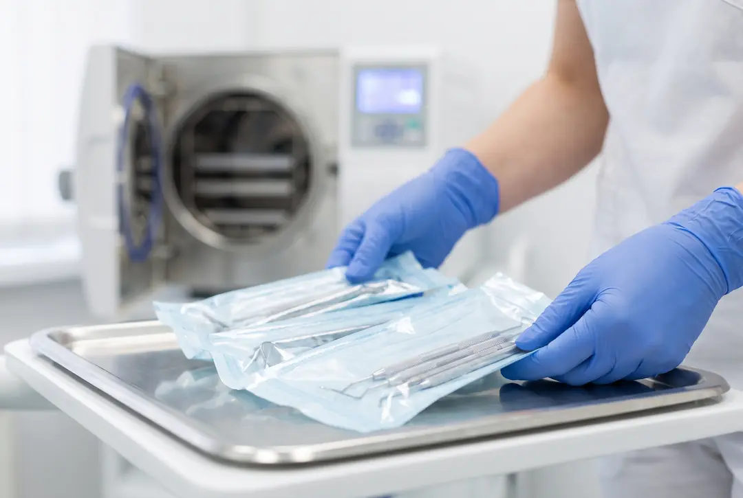 Gloved hands arranging sealed sterilisation pouches of dental instruments on a stainless steel tray